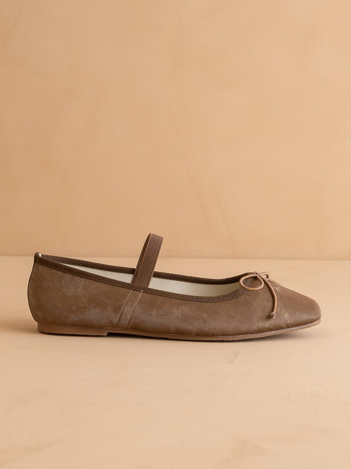 The London Ballet Flat by OASIS SOCIETY is a brown suede point flat featuring a thin elastic strap and a small bow at the toe, ideal for everyday wear and showcased on a neutral beige background.