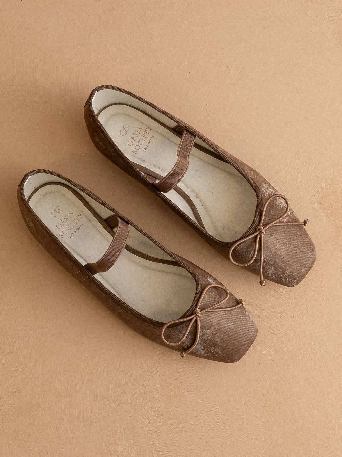 The London Ballet Flat by OASIS SOCIETY features brown square toes, bow accents, and elastic straps, making these slip-on flats perfect for everyday wear, shown on a tan surface.