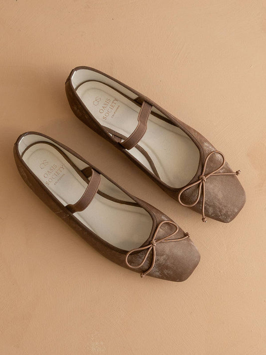The London Ballet Flat by OASIS SOCIETY features brown square toes, bow accents, and elastic straps, making these slip-on flats perfect for everyday wear, shown on a tan surface.